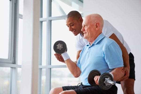 Healthy senior man exercising with dumbbells in the gym assisted by young fitness trainer